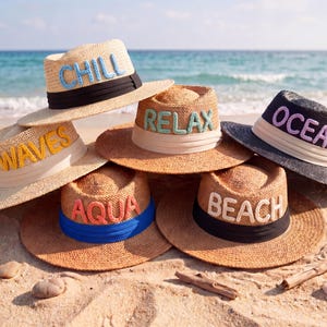 Op de afbeelding: Een verzameling strohoeden met geborduurde woorden: "CHILL", "WAVES", "RELAX", "AQUA", "BEACH" en "OCEAN". Elke hoed heeft een andere gekleurde band en letters. De hoeden liggen op een zandstrand, met de zee op de achtergrond.