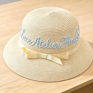 May include: A beige straw hat with a wide brim and a cream-coloured ribbon. The hat features the embroidered text "Luce Atelier Finds" in light blue script. A cream-coloured bow is tied on the front of the hat.