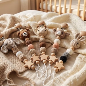 May include: Five dummy clips featuring crocheted animal heads including an elephant, bear, giraffe, deer, and rabbit. Each clip includes wooden beads, a star-shaped wooden charm, and a metal attachment, arranged in a fan shape on a textured blanket.