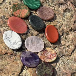 Puede incluir: Una colección de piedras preciosas pulidas de forma ovalada en varios colores, incluyendo malaquita verde, jaspe rojo y amatista púrpura. Las piedras están dispuestas sobre una superficie rocosa marrón texturizada, mostrando sus patrones y tonalidades naturales.