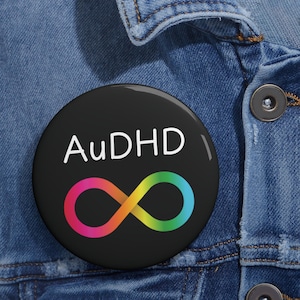 Peut inclure: Un badge rond avec un fond noir portant le texte blanc AuDHD au-dessus d'un symbole infini multicolore qui passe du rose à l'orange, au jaune et au vert, fixé sur une surface en tissu denim bleu.