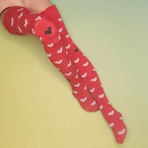 Könnte beinhalten: Rote Overknee-Socken mit weißem Herzmuster und einem schwarzen Herz am Knie. Die Socken sind aus Strickmaterial und werden vor einem hellgelben und grünen Hintergrund präsentiert.