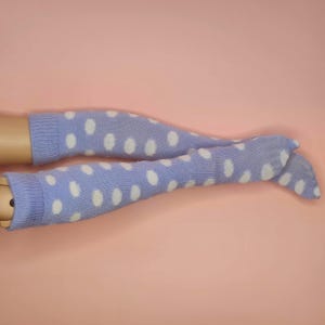 May include: A pair of light blue leg warmers with white polka dots. The leg warmers are made of a knitted material and are displayed against a pink background. The leg warmers are designed to be worn on the legs.