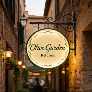 May include: A round, illuminated sign with a dark green border and decorative scrollwork, hanging from a black metal bracket. The sign features the text Olive Garden Kitchen in dark green script and serif font against a warm, glowing background.