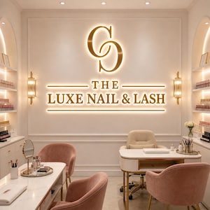 Peut inclure: Intérieur d'un salon de manucure et de cils avec un logo doré et le texte "THE LUXE NAIL & LASH" sur le mur. La pièce comprend des chaises roses, des tables blanches et des étagères exposant du vernis à ongles. L'esthétique générale est élégante et moderne.