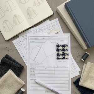 May include: A fashion design project sheet labelled Cape #0101 with a flat sketch, a blue and white houndstooth fabric swatch, and a pen. The sheet includes a table for measurements, with 85 cm written in the back length field, alongside fabric scraps and thread.