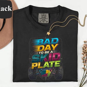 Könnte beinhalten: Schwarzes Rundhals-T-Shirt mit dem Text "BAD DAY TO BE A SKID PLATE" in einem farbenfrohen Grafikdesign. Eine goldene Halskette mit Sternanhänger liegt auf dem Shirt.