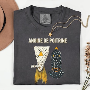 Puede incluir: Camiseta Comfort Colors gris oscuro con el texto "ANGINE DE POITRINE" y dos figuras caprichosas con forma de máscara punteada. También se ven un collar con colgante de estrella dorada y un sombrero marrón.