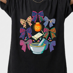 May include: Black t-shirt with a pharmacy-themed design. The graphic includes colourful bows, a pill bottle with a smiling face, a prescription, a mortar and pestle, and scattered pills. The bows have various patterns.