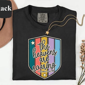 May include: Black crew neck t-shirt featuring a colourful graphic that reads "the heavens are roaring" in a shield shape. A gold star pendant necklace is draped over the shirt. The shirt is made of a soft material.