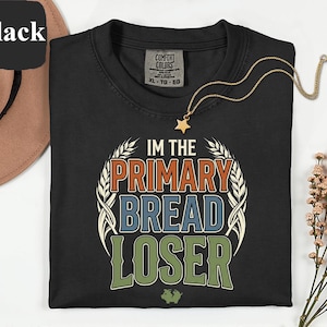 Może przedstawiać: Czarny t-shirt z okrągłym dekoltem z napisem "IM THE PRIMARY BREAD LOSER" w kolorowym wzorze. Złoty naszyjnik z wisiorkiem w kształcie gwiazdy jest zarzucony na koszulkę. Koszulka jest marki Comfort Colors.
