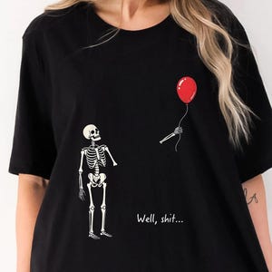 May include: A black short-sleeved t-shirt featuring a graphic of a white skeleton looking up at a red balloon held by a detached skeletal hand. The text "Well, shit..." is printed in white below the skeleton on the front of the garment.