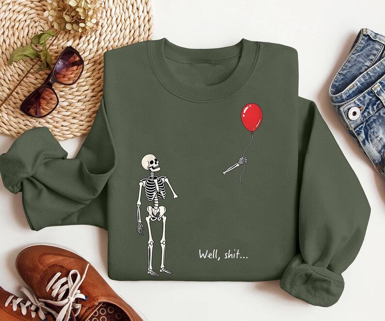 May include: An olive green crewneck sweatshirt featuring a graphic print of a skeleton looking up at a detached skeletal hand holding a red balloon. The text "Well, shit..." is printed in white below the skeleton on the front of the garment.