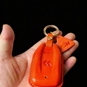 Puede incluir: Llavero de cuero naranja con un llavero dorado y una pequeña etiqueta naranja. El llavero tiene un borde cosido y un logotipo en relieve. La etiqueta presenta un logotipo "M" en relieve. El llavero se sostiene en una mano sobre un fondo negro.