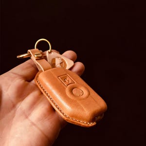 May include: A tan leather key fob with a gold-colored key ring and a matching leather tag. The key fob has a stitched border and a raised design. The background is dark.