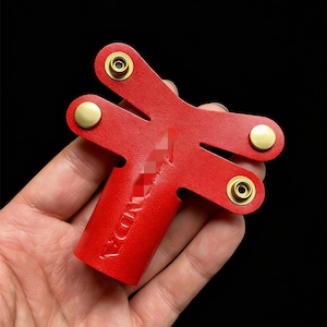 May include: A red leather key organizer with a Y-shaped design. The organizer has three sections and is secured with brass rivets. The word "VANDA" is embossed on the side. The background is black.
