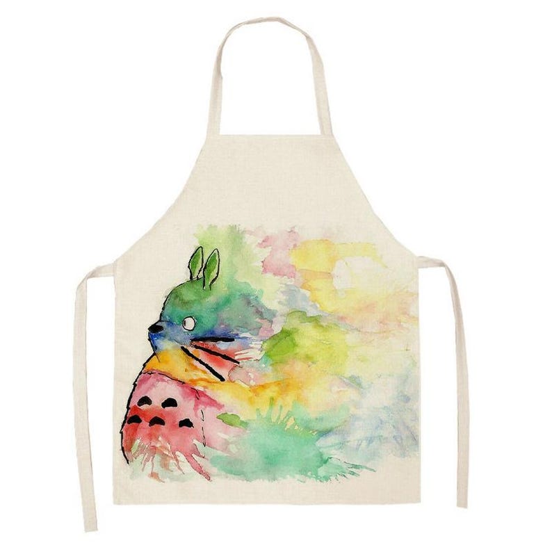 Puede incluir: Un delantal color crema con una ilustraci&oacute;n de acuarela de un personaje de dibujos animados. El personaje es principalmente verde, azul y rosa, con un dise&ntilde;o caprichoso. El delantal tiene un lazo para el cuello y lazos en la cintura.