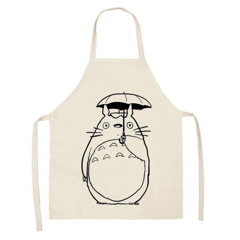 Puede incluir: Delantal beige con una ilustraci&oacute;n en blanco y negro de un personaje de dibujos animados sosteniendo un paraguas. El delantal tiene un lazo para el cuello y lazos en la cintura, adecuado para cocinar o manualidades.