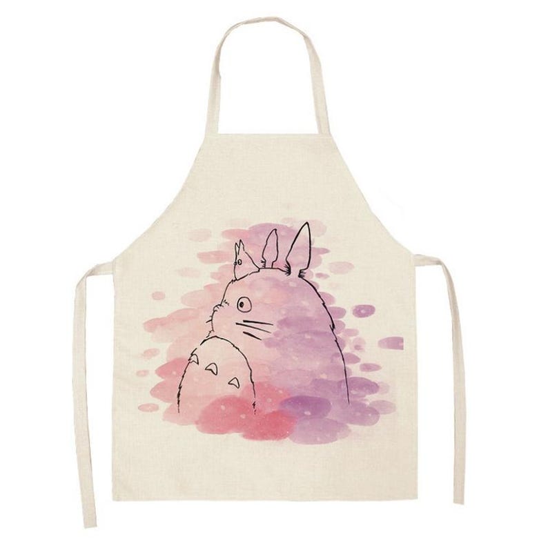 Puede incluir: Un delantal beige con una ilustraci&oacute;n de acuarela de dos personajes de dibujos animados en tonos morados y rosas. El delantal tiene un lazo para el cuello y lazos en la cintura. El dise&ntilde;o es de estilo caprichoso y art&iacute;stico.