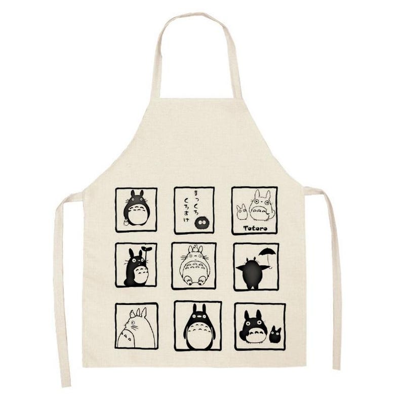 Puede incluir: Delantal beige con ilustraciones en blanco y negro de personajes de Totoro en marcos cuadrados. El delantal tiene un lazo para el cuello y lazos en la cintura. La palabra "Totoro" est&aacute; impresa en el delantal.