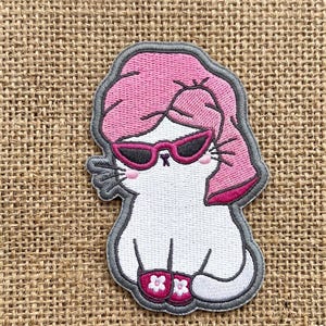 May include: Embroidered patch featuring a white cat with pink hair and sunglasses. The cat is wearing pink flower slippers and has a gray border. The patch is designed to be ironed on or sewn onto fabric.