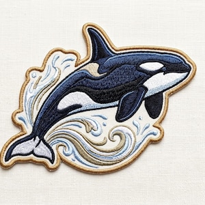 May include: Embroidered patch featuring a detailed orca whale leaping through stylized waves. The whale is primarily navy blue and white, with black accents. The patch has a gold border and is set against a cream-colored background.
