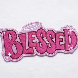 May include: A pink embroidered patch with the word "BLESSED" in a bubble letter font. The patch has a white halo above the word and white starburst accents. The patch is on a white background.