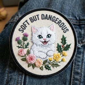 May include: Embroidered patch with a white cat illustration, surrounded by flowers and greenery. The text "SOFT BUT DANGEROUS" is arched around the top. The cat has green eyes and a slightly open mouth, revealing teeth.