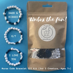 Op de afbeelding: Een Morsecode armband DIY kit met een bruine papieren zak, een doorzichtig venster en drie afgewerkte armbanden. De armbanden zijn witte en zwarte kralen die "HAGS & KISSES", "6/7" en "I LOVE YOU" spellen. Op de zak staat "Unbox the fun!"