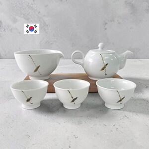 Puede incluir: Un juego de té de cerámica blanca que consta de una tetera, una jarra vertedora y tres tazas de té pequeñas. Cada pieza presenta un delicado motivo botánico marrón pintado a mano. El conjunto está dispuesto sobre una base de madera rectangular.
