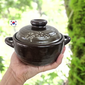 以下が含まれることがあります： 蓋付きのダークブラウンの陶器製ポットで、両側にハンドルが付いています。蓋には淡い色のアクセントが入った繊細な花のエンボス模様が施されています。ポットは光沢のある滑らかな仕上げで、深みのあるボウル型をしています。