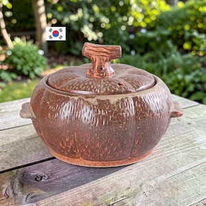 Puede incluir: Una olla de cerámica marrón con tapa y una superficie texturizada y acanalada que recuerda a una calabaza. El recipiente cuenta con dos pequeñas asas laterales y un asa en forma de T en la tapa. El esmalte tiene un acabado terroso y moteado.