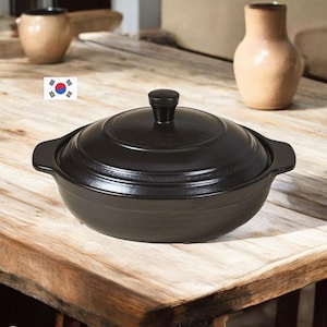 Puede incluir: Una olla de cocina de cerámica negra con tapa a juego y dos asas laterales. La olla tiene un acabado oscuro y liso, con un pomo central en la tapa. Se puede ver un pequeño icono de la bandera de Corea del Sur en la imagen.