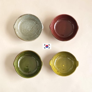 May include: Four round ceramic bowls with small side handles arranged in a square formation. The bowls feature distinct glossy glazes in light gray with a crackle finish, deep maroon, dark olive green, and a vibrant yellow-green color.