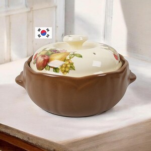 May include: A brown ceramic cooking pot with a cream-colored lid featuring a painted fruit motif of a red apple, a yellow pear, and green grapes. The pot has two side handles and a central knob on the lid, which includes a small steam vent hole.