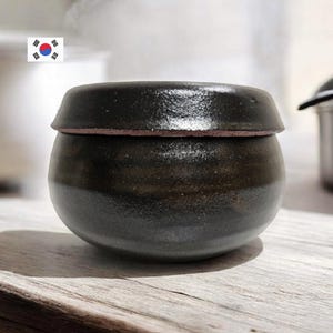 Puede incluir: Un tarro de cerámica marrón oscuro con tapa. El tarro tiene un cuerpo redondeado y un borde ligeramente ensanchado. La tapa es plana y encaja perfectamente. El tarro parece hecho a mano y tiene una estética rústica y terrosa.