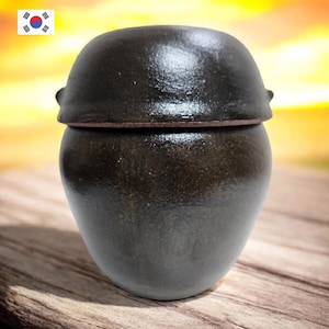 Peut inclure: Un pot en céramique brun foncé avec un couvercle. Le pot a un corps arrondi et un couvercle légèrement bombé. Le pot est posé sur une surface en bois. L'arrière-plan est flou, avec des teintes jaunes et orangées.