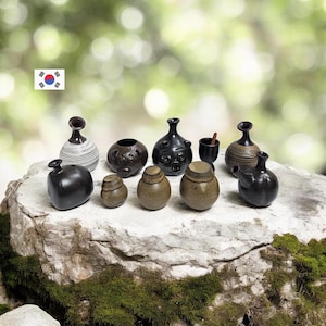 May include: A collection of handcrafted ceramic vessels in various shapes and sizes. The pottery includes small jars, vases, and a cup, with colors ranging from black and brown to tan and white. The items are displayed on a stone surface.
