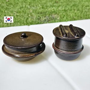 May include: Two brown ceramic cooking pots with lids. One pot has a round lid with a small knob, and the other has a lid with two handles. The pots are on a white surface with a green background.