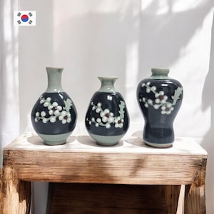 May include: Three ceramic vases with a dark blue glaze and white floral designs. Each vase has a light green base and rim. The vases are displayed on a wooden surface, showcasing a blend of traditional and modern aesthetics.