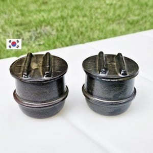 May include: Two dark gray ceramic containers with lids. Each container has a rounded base and a lid with two raised handles. The containers are set against a background of green grass and a white surface.