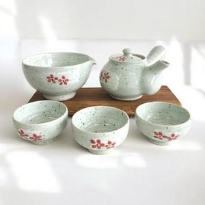 May include: A ceramic tea set featuring a light green speckled glaze with hand-painted red flower motifs. The set includes one teapot with a side handle, one large pouring bowl with a spout, and three small matching tea cups.