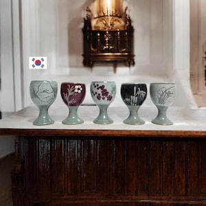 May include: Five ceramic goblets with unique designs. The goblets have a light green stem and base, with a variety of painted designs on the bowl, including floral and bamboo motifs. The goblets are arranged in a row on a wooden surface.