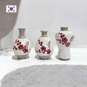 May include: Three ceramic vases with a cream-colored base and gray tops. Each vase is adorned with a red floral design. The vases vary in shape, with one being more rounded and another taller and more slender.