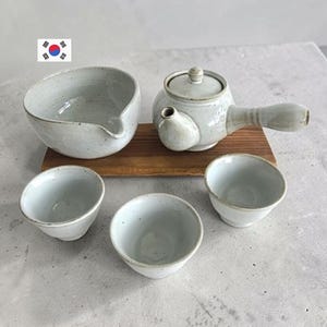 Op de afbeelding: Een keramische theeset bestaande uit een theepot met zijhandvat, een schenkkom en drie kleine kopjes, allemaal afgewerkt met een gespikkeld gebroken wit glazuur. De theepot en schenkkom staan op een rechthoekig houten dienblad.