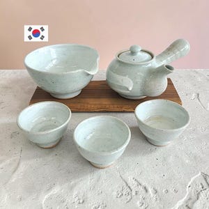 Może przedstawiać: Ceramiczny zestaw do herbaty z jasnoniebiesko-szarą nakrapianą glazurą. Zestaw zawiera czajniczek z bocznym uchwytem i pokrywką, miseczkę do nalewania oraz trzy małe filiżanki do herbaty, ustawione na prostokątnej drewnianej desce.