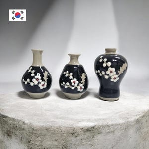 May include: Three ceramic vases with a dark blue glaze and floral designs. The vases have a cream-colored neck and base. The floral design features white flowers with red centers. The vases are of varying shapes and sizes.