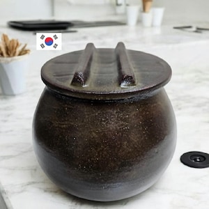 May include: A dark brown ceramic jar with a lid. The jar has a rounded body and a lid with two raised handles. The jar is sitting on a white marble surface. A small black object is to the right of the jar.