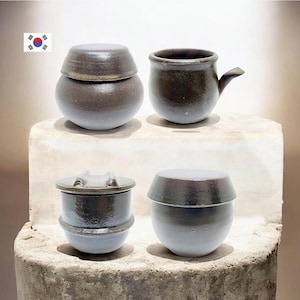 Puede incluir: Cuatro recipientes de cerámica hechos a mano de varias formas y tamaños. La cerámica presenta una mezcla de esmaltes marrón oscuro y gris claro. Una pieza tiene un asa. Las piezas se exhiben sobre una superficie texturizada de color gris claro.