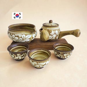 Puede incluir: Un juego de té de cerámica que incluye una tetera con mango lateral, un cuenco vertedor y tres tazas pequeñas. Cada pieza tiene un esmalte marrón moteado con un patrón de hojas blancas pintadas a mano sobre una base de madera.